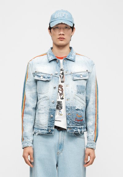 Άνδρας που φοράει ανοιχτό μπλε τζιν μπουφάν με φθαρμένες λεπτομέρειες και ρίγες ουράνιου τόξου στα μανίκια, σετ με τζιν παντελόνι και τζιν καπέλο με το κείμενο "HOT".