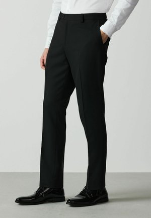 Mann trägt schwarze maßgeschneiderte Anzughose, weißes Hemd und polierte schwarze Anzugsschuhe und steht mit einer Hand in der Hosentasche.