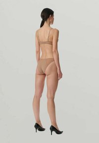 Donna in piedi di spalle che indossa un reggiseno e delle mutande beige con tacchi neri su uno sfondo chiaro e semplice.