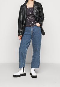 Blazer de acrílico negro sobre una blusa sin mangas de patrón oscuro, combinado con unos vaqueros de pierna ancha de tiro alto en azul y botas blancas hasta el tobillo.
