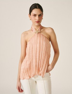 Femeie purtând un top piersiciu plisat cu decolteu halter, decorat cu o broșă în formă de stea de mare, și pantaloni albi, stând în fața unui fundal simplu și deschis la culoare.