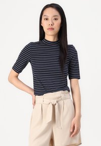 Tricou scurt cu mâneci scurte, în dungi bleumarin și alb, cu guler înalt, asortat cu pantaloni scurți bej, cu talie înaltă, având detaliu cu fundă la talie.