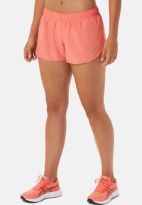 Piernas femeninas vestidas con pantalones cortos deportivos color coral y zapatillas de correr a juego, de pie contra un fondo blanco.