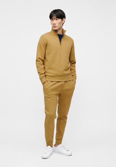 Άνδρας στέκεται με τα χέρια στις τσέπες, φορώντας ζιπ-πουλόβερ καμηλό χρώματος και παντελόνι τζόγκινγκ, συνδυασμένα με λευκά αθλητικά παπούτσια, πάνω σε απλό λευκό φόντο.