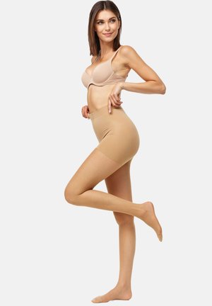 Pantalones cortos moldeadores del color de la piel con una textura suave, cintura alta y longitud a mitad del muslo, la modelo posa con una pierna levantada, vistiendo un sujetador a juego.