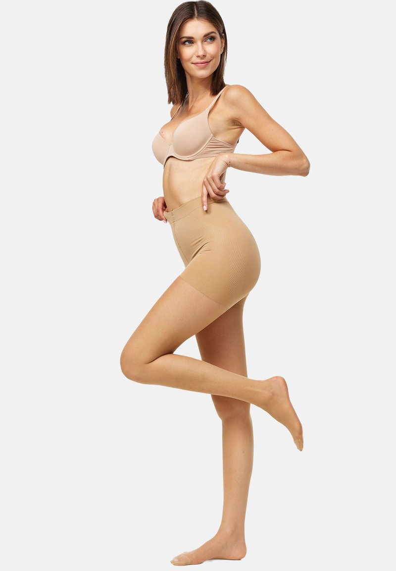 Pantalones cortos moldeadores del color de la piel con una textura suave, cintura alta y longitud a mitad del muslo, la modelo posa con una pierna levantada, vistiendo un sujetador a juego.