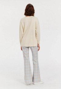 Pull en maille beige à manches longues et coupe décontractée, associé à un pantalon à carreaux bleu clair évasé avec des accents jaunes. Des baskets blanches sont visibles.