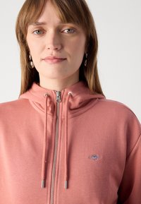 Mikina na zips v svetloružovej farbe z mäkkého materiálu, s metalickými prvkami, nastaviteľnými šnúrkami a malým vyšívaným logom.