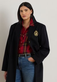 Black wool coat with a crest on the left chest, paired with a red and yellow plaid shirt. Worn with blue jeans and a black belt.