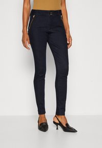 Jeans skinny de mezclilla azul oscuro con bolsillos con cremallera, cierre de dos botones y una textura suave. Usados con zapatos de tacón negros.