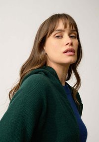 Zielony kardigan z kapturem, wykonany z miękkiego materiału, noszony na niebieskiej bluzie z okrągłym dekoltem. Charakteryzuje się luźnym krojem i minimalistycznym stylem.