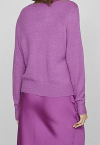 Fialový pletený svetr s dlouhými rukávy a žebrovanými manžetami. Měkká textura a volný střih, sladěný s odpovídající saténovou sukní, také fialovou.