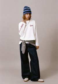 Maglietta bianca a maniche lunghe con scritte rosse, maglione grigio legato in vita, jeans a gamba larga scuri, berretto a righe blu e grigio, scarpe da ginnastica bianche.