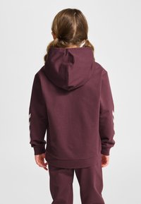 Bordowa bluza z kapturem z miękkiego materiału, z dużym kapturem, ściągaczami na mankietach oraz białymi ukośnymi paskami na rękawach. Noszona wraz z pasującymi spodniami.