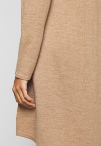 Vêtement en tricot beige clair avec une finition texturée. Présente un ourlet légèrement évasé et des poignets ajustés, avec une main positionnée près du bord.