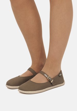 Piedi che indossano scarpe piatte stile Mary Jane marroni con cinturino regolabile e suole bianche su sfondo chiaro.