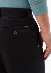Pantaloni in denim nero con dettagli sulla tasca posteriore, bottone argento e etichetta con logo. Texture liscia, design a gamba dritta, vista ravvicinata.