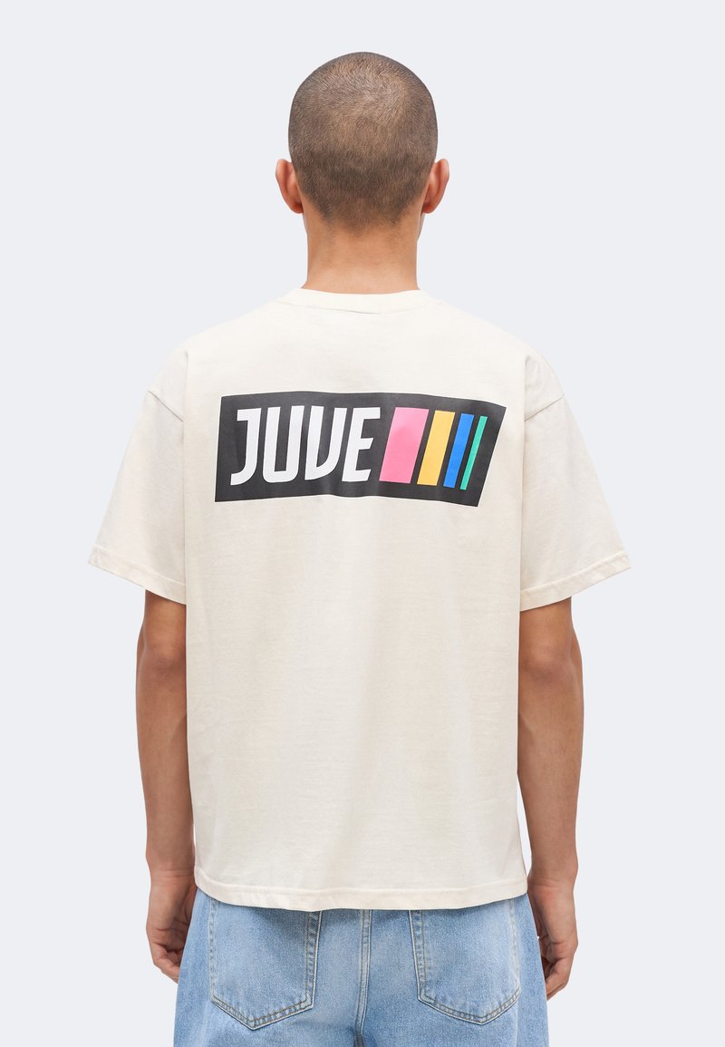 Άνδρας με κοντά μαλλιά που φοράει λευκό T-shirt με τη λέξη «JUVE» και ροζ, κίτρινες, μπλε ρίγες στην πλάτη, συνδυασμένο με ανοιχτό μπλε τζιν.