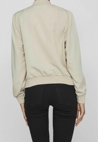 Personne portant une veste beige à manches longues et un pantalon noir ajusté, debout face à un fond gris clair.