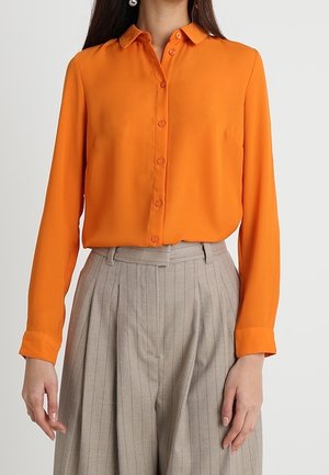 Mujer con una blusa naranja de manga larga y botones, metida dentro de unos pantalones de pinzas beige de talle alto con rayas finas.