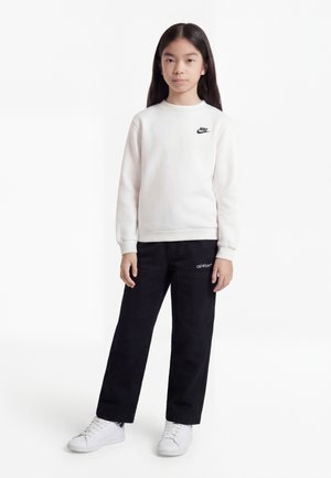 Niño con cabello largo y oscuro que lleva una sudadera blanca de Nike, pantalones negros de Off-White y zapatillas deportivas blancas, posando contra un fondo blanco.