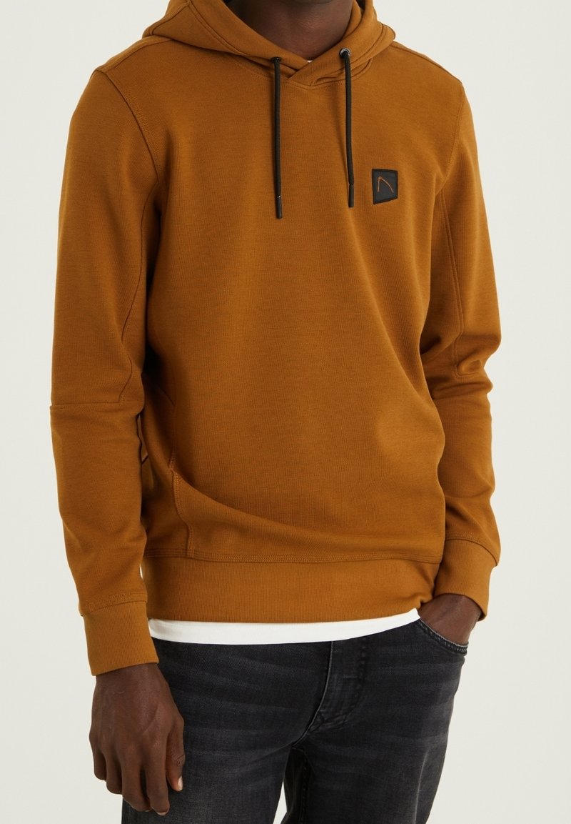 Homme portant un sweat à capuche couleur moutarde avec des cordons noirs et un petit patch noir sur la poitrine, associé à un jean noir et une chemise blanche en dessous.