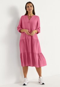 Rosa gestuftes Kleid mit langen Ärmeln, rundem Ausschnitt undBindedetail. Weicher Stoff, lockere Passform und knielanges Design. Getragen mit weißen Sneakers.
