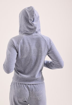 Persona che indossa una tuta grigia con cappuccio con la scritta "Juicy" in strass sulla schiena della giacca e sui pantaloni, vista da dietro.