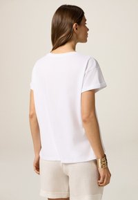 Camisa de algodón blanca con mangas cortas enrolladas, un cuello redondo y un ajuste relajado, vista desde la parte de atrás. Textura simple y suave.