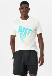 Bela bombažna majica z velikim modrim in bež grafičnim tiskom, ki pravi "SIMPLE NAREDITE". Nosena z črnimi kratkimi hlačami, ustreza bež ozadju.