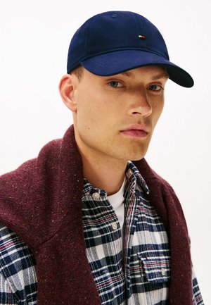 Navy cap with a small multicolored logo, worn with a maroon sweater over a blue and white checkered flannel shirt. Textured fabric visible.
