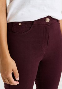 Jeans ajustados burdeos con una textura suave, que cuentan con un bolsillo delantero, cierre de botón de metal y acentos de costura. La mano del modelo está en la cintura.