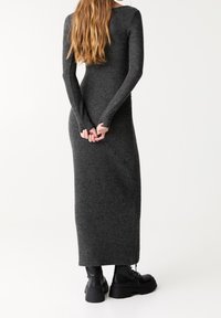 Enganliegendes graues Kleid mit langen Ärmeln aus weichem Strickmaterial, mit gerader Silhouette und seitlichem Schlitz, kombiniert mit klobigen schwarzen Stiefeln.