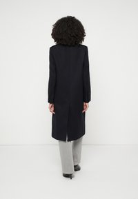 Persoană cu păr creț, purtând un palton lung negru, pantaloni gri deschis și pantofi cu toc negri, stând în fața unui fundal alb, cu fața întoarsă.