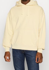 Sudadera amarilla claro con un bolsillo frontal, cordones ajustables y puños de canalé. La tela es suave, con un corte relajado y un branding mínimo.