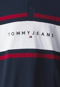 Polo navy con rayas horizontales blancas y rojas. Presenta un collar y el texto "TOMMYJEANS" bordado en negro. Logo en rojo, blanco y azul.