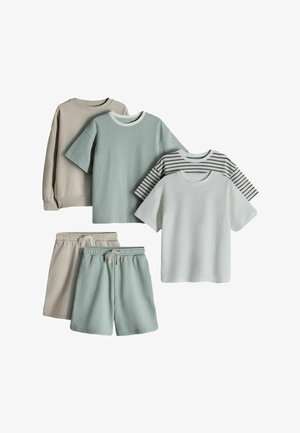 Smilende barn med skulderlangt hår iført en stribet skjorte og lyse shorts, der holder en lyse sweater bag ryggen.