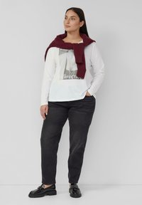 Witte lange mouwen shirt met een grafische print, zwarte spijkerbroek en zwarte lakleren loafers. Een wijnrood trui is over de schouders gedrapeerd.