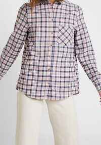 Rutig flanellskjorta i ljusrosa, marinblå och vit med långa ärmar, knappframsida och en enkel bröstficka med rutigt mönster.