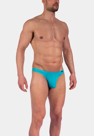 Muskuløs mand iført lyseblå lavtaljede underbukser, stående med armene afslappet ned langs siderne mod en ensartet lys grå baggrund.