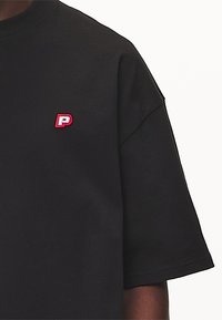 Černé bavlněné tričko s krátkým rukávem, na levé části hrudi s malým vyšitým logem "P" v červené barvě. Hladká textura.