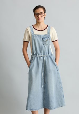 Młoda kobieta w okularach, kremowym t-shircie i jasnoniebieskiej sukience ogrodniczce z dżinsu, z rękami w kieszeniach, stojąca na gładkim tle.