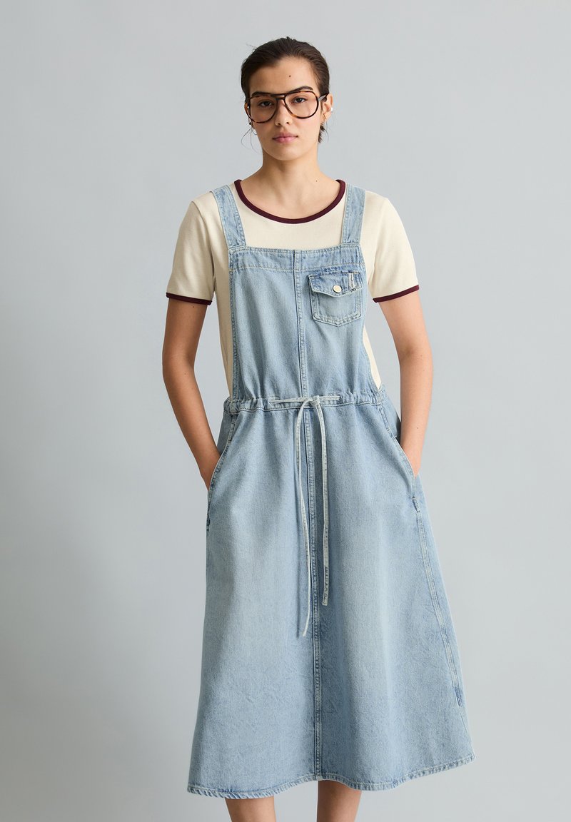 Junge Frau mit Brille, cremefarbenem T-Shirt und hellblauem Jeans-Overallkleid, die mit den Händen in den Taschen vor einem einfarbigen Hintergrund steht.