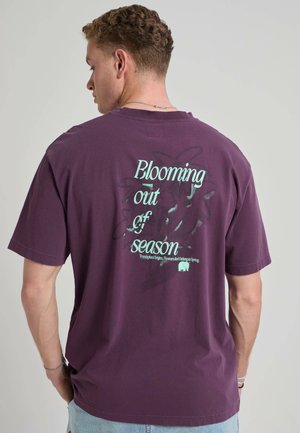 Man draagt een paars t-shirt met blauwe tekst "Blooming out of season" en subtiele bloemmotieven op de rug, tegen een effen achtergrond.