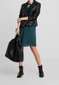 Chaqueta de motociclista de cuero negro sobre un vestido verde, llevando un bolso de mimbre negro. Botines completan el conjunto, que presentan detalles texturizados.