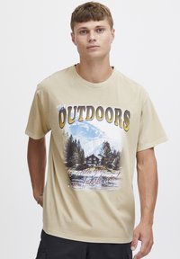 Beige bomulls T-skjorte med et landskapsgrafikk av fjell, en hytte og fugler. Teksten sier "UTENDØRS" med fete farger.
