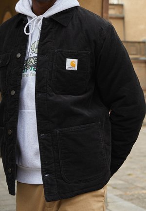 Mężczyzna w czarnej sztruksowej kurtce Carhartt na jasnoszarej bluzie z kapturem z zielonym napisem i beżowych spodniach, stojący na zewnątrz na chodniku.