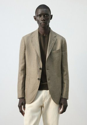 Schwarzer Mann trägt ein beiges karriertes Sakko über einem braunen Hemd und cremefarbenen Hosen, steht vor einem schlichten hellen Hintergrund.