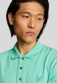 Camisa polo verde menta con un tejido texturizado, cuello corto y tres botones oscuros. Presenta un pequeño logo bordado en el pecho.