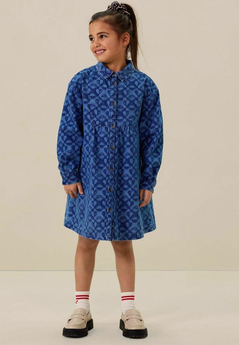 Robe en denim bleu avec un motif, devant à boutons, et manches longues ; portée avec des chaussettes blanches, des accents rouges et des chaussures beiges.